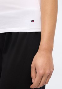 Λευκό τελείωμα πουκάμισου με μικρό λογότυπο Tommy Hilfiger πάνω από μαύρο παντελόνι, και χαλαρό δεξί χέρι δίπλα στο σώμα.