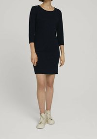Mörkblå stickad klänning med trekvartslånga ärmar, figurnära design, rund halsringning, i kombination med ljusbeige hög-top sneakers. Slät textur.
