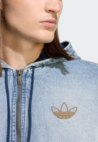 Farmer anyagú kapucnis felső cipzáras elejével, kék belső béléssel és arany hímzett logóval a mellkason. Sima tapintású és laza, kényelmes szabású.