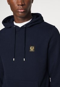 Jūras zila kapuce no mīksta auduma ar priekšējo kabatu, ar taisnstūra zelta logo uzļaipus un regulējamiem aukliņām ar metāla galiem.