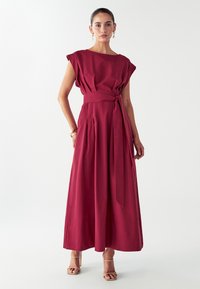 Bordový maxi šaty s krátkymi dolman rukávmi, opaskom v páse a skladanými detailmi. Mäkka textúra látky a splývavý strih.