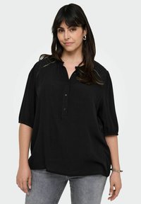 Blusa nera con una fila di bottoni e maniche a palloncino corte. Presenta dettagli in pizzo sulle spalle. Tessuto morbido, vestibilità ampia, abbinata a jeans grigi.