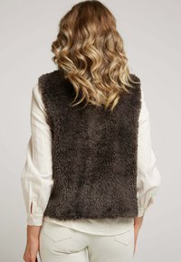 Gilet en polaire marron avec col haut et sans manches, présentant une surface douce et texturée. Port é par-dessus une chemise à manches longues crème et un pantalon blanc.