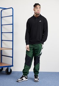Pólo preto de mangas longas, calças de treino verde e pretas com detalhes em malha, e ténis pretos e brancos. Cores sólidas com detalhes de logótipos.