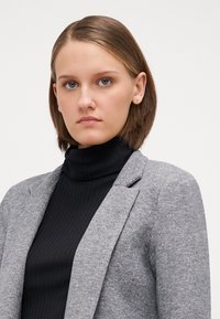 Siv blazer z teksturirano tkanino, z detajli na reverju, nosi se preko črnega rebrastega vratnega puloverja. Ravni, do ramen dolgi lasje. Nevtralna ozadja.