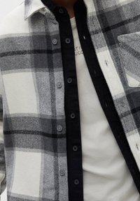 Szary i biały koszula flanelowa w kratę z zapięciem na guziki, czarnymi akcentami i kieszonką na piersi, noszona na jasnoszarej koszulce.