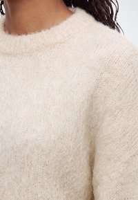 Gros plan d'une personne portant un pull en maille doux et duveteux de couleur crème avec un col rond.