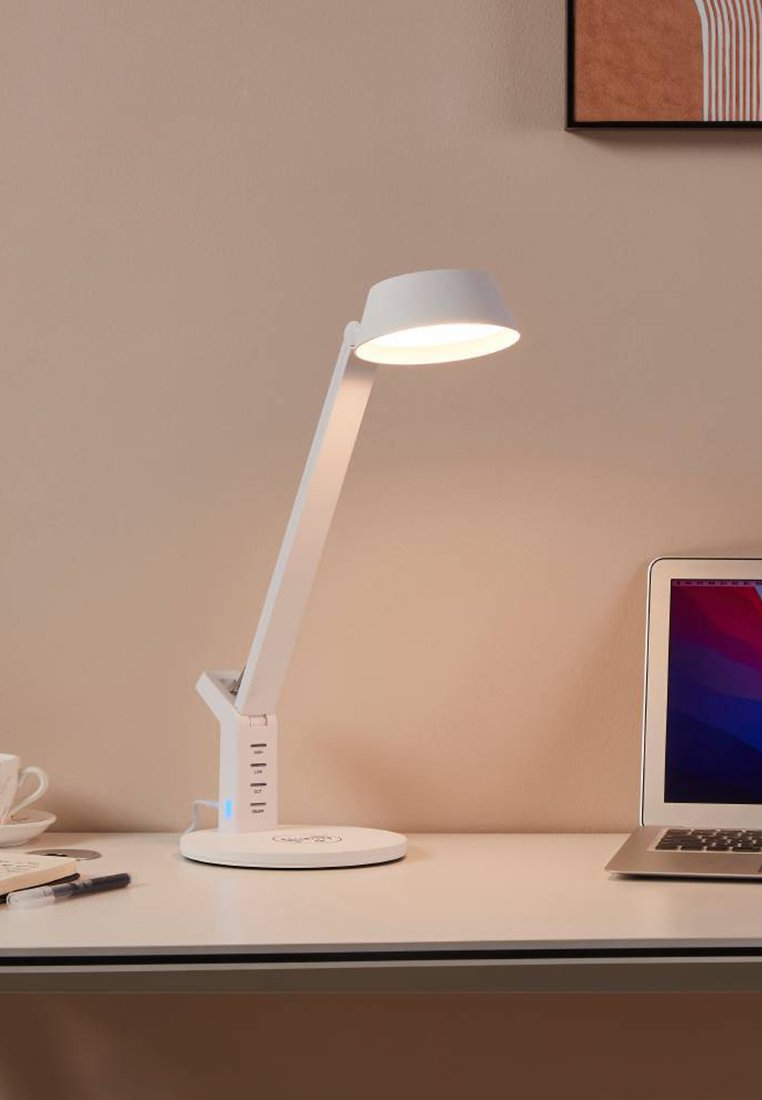 Biała regulowana lampa biurkowa z wbudowanymi portami USB oświetla miejsce pracy z laptopem, kubkiem, spodeczkiem, notatnikiem i długopisem na beżowej powierzchni i ścianie.