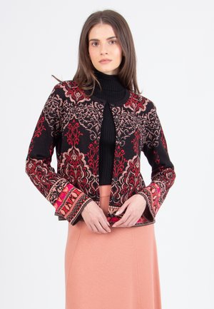 Vrouw in een zwarte coltrui, een gestreepte zwart-rode cardigan met knopen, en een perzikkleurige rok, staand tegen een witte achtergrond.