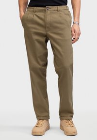 Man wearing olive green chinos, black shirt, beige sneakers, with hands in pockets, standing against plain light background.