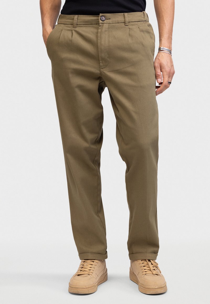 Man wearing olive green chinos, black shirt, beige sneakers, with hands in pockets, standing against plain light background.