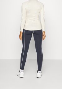 Krátký rukáv sportovní top v krémové barvě s geometrickým vzorem a kontrastním černým logem, doplněný o tmavě modré legíny s bílými bočními pruhy.