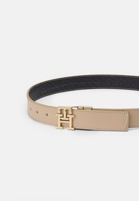 Ceinture en cuir beige avec une boucle en métal doré au ton chaud, ornée d'un logo "H" entrelacé. Intérieur noir à motifs avec des coutures contrastantes.