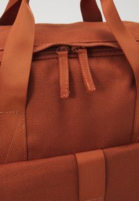 Sac en toile couleur rouille avec deux curseurs de fermeture éclair sur le dessus, anses renforcées et bretelles cousues pour le transport.