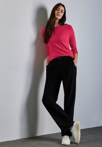 Maglietta rosa a maniche lunghe aderente abbinata a pantaloni neri su misura, con tasche laterali e sneakers bianche, su uno sfondo grigio chiaro.