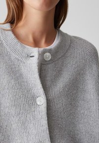 Gros plan sur une personne portant un cardigan boutonné en tissu texturé de couleur gris clair sur un haut en jersey blanc, mettant en évidence les détails du cou et du haut de la poitrine.