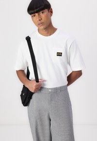 Tricou alb cu mânecă scurtă, având un buzunar pe piept cu o etichetă neagră. Modelul poartă pantaloni rai și ține o geantă mică neagră cu un breloc.