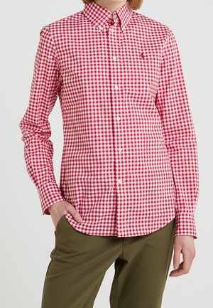 Personne portant une chemise à carreaux rouge et blanche boutonnée et un pantalon vert olive, une main dans la poche, sur un fond uni.