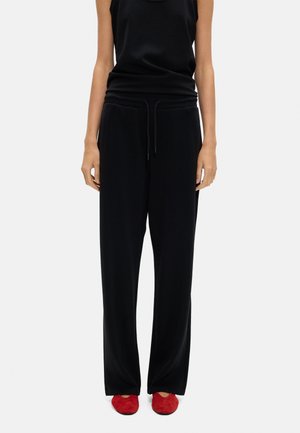 Osoba ubrana w czarne luźne spodnie i czarną bezrękawnikową bluzkę, w połączeniu z czerwonymi płaskimi butami, stojąca na jednolitym tle.