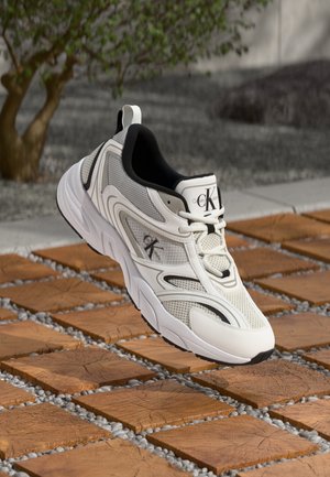 Biało-szary sneaker Calvin Klein spoczywający na drewnianej płytce i żwirowym podłożu z zamazanym drzewem w tle.