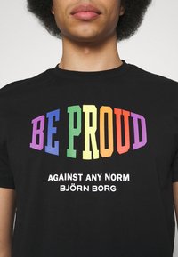 Czarny t-shirt z wyrazistym, wielokolorowym napisem "BE PROUD" oraz białym napisem poniżej "AGAINST ANY NORM BJÖRN BORG." Standardowy design z okrągłym dekoltem.