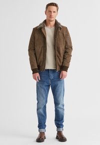 Braune Wildleder-Bomberjacke mit einem Fellkragen, zwei seitlichen Taschen, gerippten Bündchen und einer mit Reißverschluss versehenen Tasche am Ärmel. Kombiniert mit blauen Jeans und braunen Stiefeln.