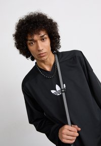 Musta, tekstuurilt sporditopp V-kaelusega, millel on valge Adidas'i logo. Mudel hoiab diagonaalselt triibulist rihma.