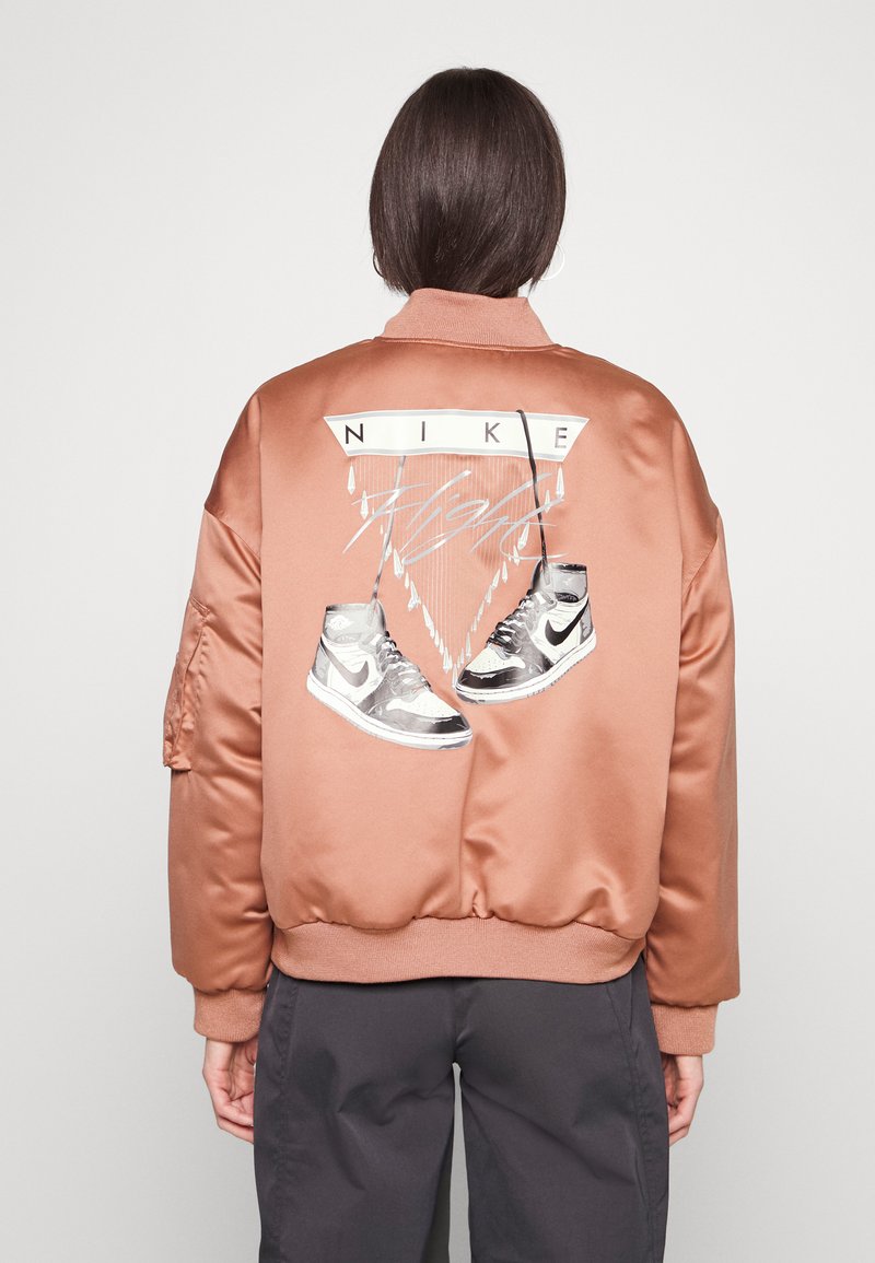 Veste bomber en satin bronze avec le texte blanc "NIKE" et des graphiques de baskets argentés au dos, dotée de poignets et d'un ourlet à côtes.