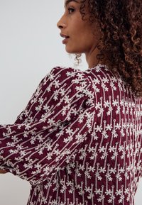 Burgundia punane kangast topp, millel on keerukas valge lilleline tikand. Disainis on tekstuuriga padjandusega varrukad ja struktureeritud kuju.