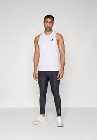 Biely tielkový top bez rukávov s čiernym logom Adidas, v kombinácii s priliehavými čiernymi legínami a bielymi športovými topánkami, na neutrálnej pozadí.