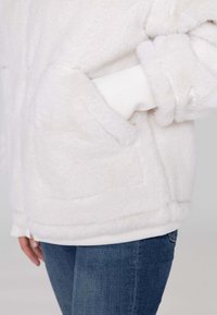 Fluffy witte jas met ribbels aan de mouwen en onderrand, voorzien van een voorzak. Gecombineerd met denim jeans, die een zachte textuur en een relaxed fit laten zien.