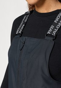 Fekete mellény állítható pántokkal, amelyen a "Salomon" felirat található. Az anyag könnyű, elöl cipzáros és texturált.