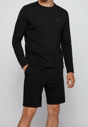 Mand iført en ensfarvet sort langærmet trøje og sorte shorts, stående med den ene hånd i lommen mod en lysegrå baggrund.
