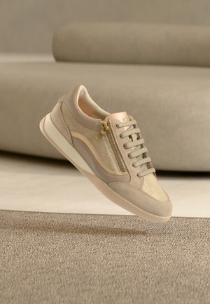 Zapatilla casual beige con cremallera dorada, cordones y suela de goma, mostrada flotando sobre una alfombra texturizada y un fondo con sofá beige.