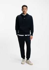 Navy hooded sweatshirt with front pocket and drawstring, layered over a white collared shirt; paired with matching joggers and beige sneakers.
