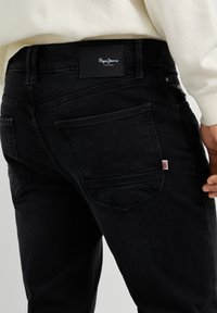 Osoba nosiaca čierne Pepe Jeans s viditeľnou zadnou vreckovou a logom, oblečená v svetlej dlhoruká blúzka.