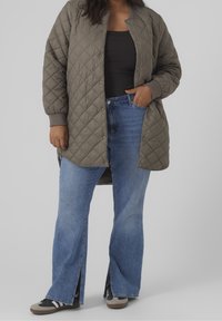 Giacca lunga imbottita in verde oliva con polsini a costine, abbinata a jeans blu a zampa con spacchi laterali e sneakers nere.