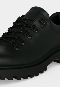 Chaussure de randonnée en cuir noir avec une semelle épaisse à crampons, un bout rond et des lacets noirs passant dans des œillets et crochets métalliques.