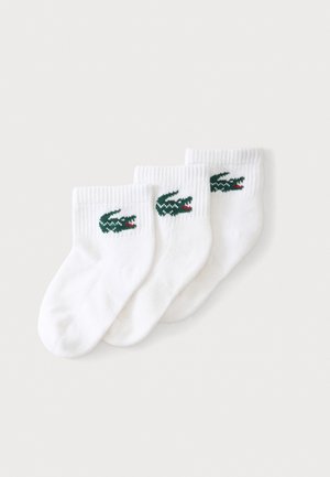 Trois paires de chaussettes blanches chevilles avec des logos de crocodile verts sur les revers, disposées en rangée sur un fond clair.