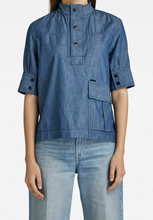 Blauw denim overhemd met korte mouwen, hoge kraag, knoopsluiting, knopen aan de manchetten en een groot klepzakje, gedragen met lichtblauwe spijkerbroek.