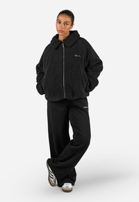 Giacca nera in pile con zip e ampio colletto, abbinata a pantaloni neri larghi. Presenta un logo sul petto della giacca. Design strutturato e casual.