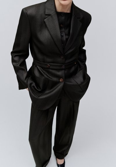 Personne portant un costume noir sur mesure avec un blazer ceinturé, une chemise boutonnée, un pantalon ample et des chaussures noires, debout les mains dans les poches.