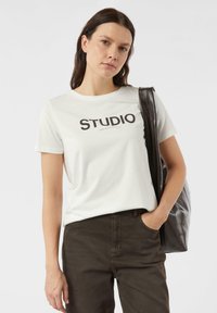 Biała koszulka z czarnym napisem "STUDIO", krótkie rękawy, miękka bawełniana tkanina, w połączeniu z ciemnobrązowymi spodniami i czarną torbą na ramię.