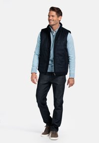 Dunkelblaues Steppwesten über einem hellblauen Langarmshirt, kombiniert mit dunklen Jeans und grauen Sneakers. Glattes Material, Reißverschluss, lässige Passform.