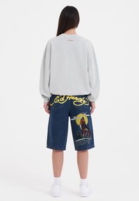Grå oversized sweatshirt med broderad logotyp; blå denimshorts med en vargdesign och gula accenter; vita sneakers.