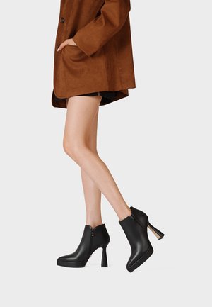 Persona che indossa un cappotto marrone e stivaletti neri con tacchi alti, mostrando le gambe incrociate su uno sfondo chiaro e uniforme.