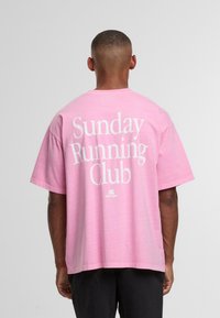 Mees seljaga, kandes lõhnavat roosa T-särki, mille seljal on valged tähestikud "Pühapäeva jooksugrupp", koos mustade pükse.