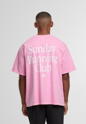Man som står med ryggen mot kameran iförd en lös rosa t-shirt med texten "Sunday Running Club" i vitt på ryggen, tillsammans med svarta byxor.