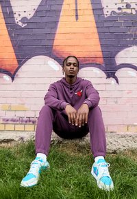 Sudadera morada y pantalones de chándal con un logo rojo, combinados con zapatillas blancas y azules. Sentado contra una pared de graffiti colorida. Césped en primer plano.