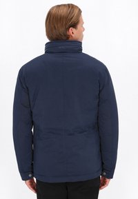 Mørkeblå pufferjakke med høj krave og knaplukning ved håndleddene; glat tekstur, minimal design og syninger der løber lodret på ryggen.